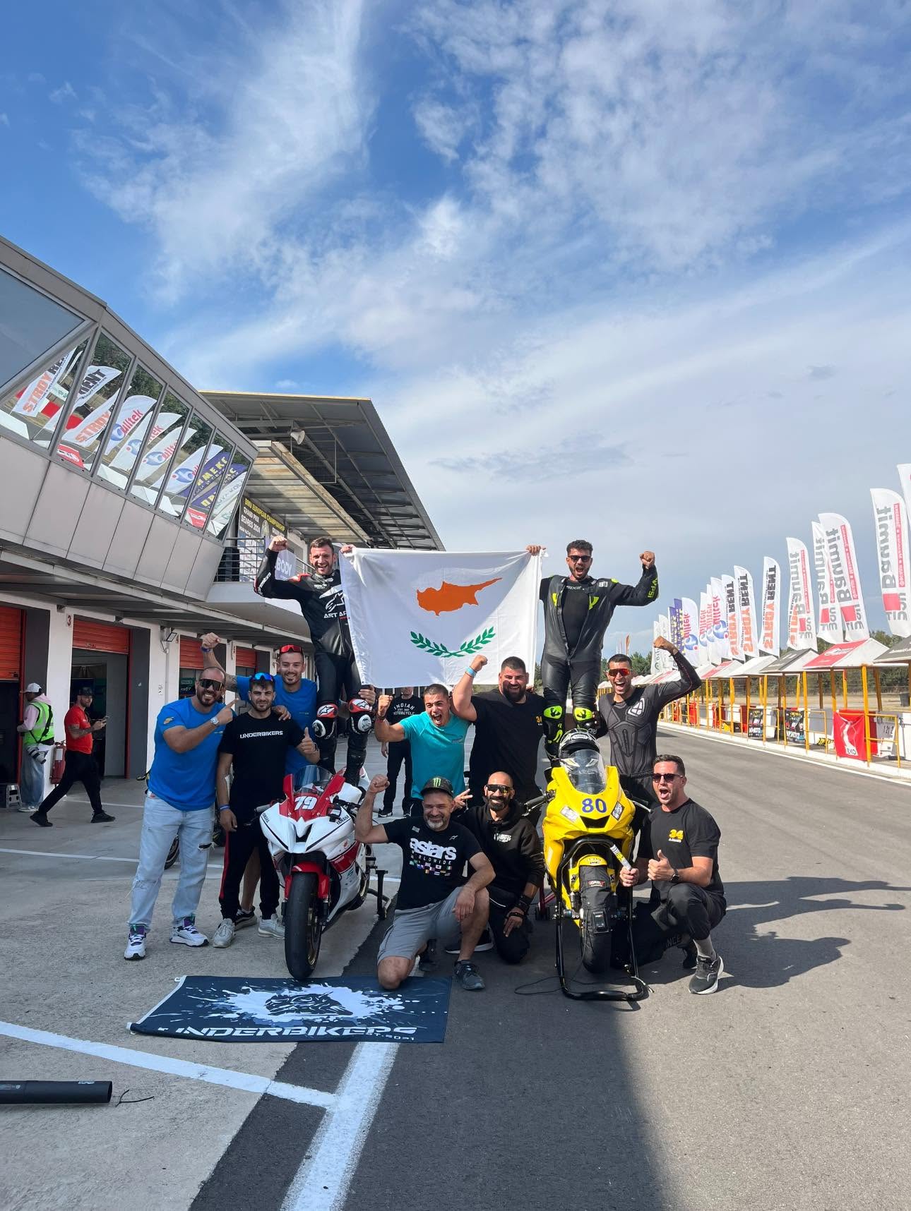 Ομάδα αναβατών και φίλων στο pit lane πίσω από μια σημαία της Κύπρου, με δύο αγωνιστικές μοτοσυκλέτες μπροστά